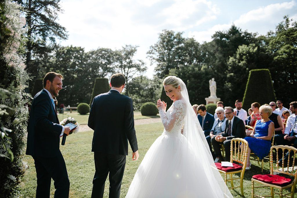Bride in lace gown waves to guests during elegant outdoor wedding ceremony on manicured lawn.
