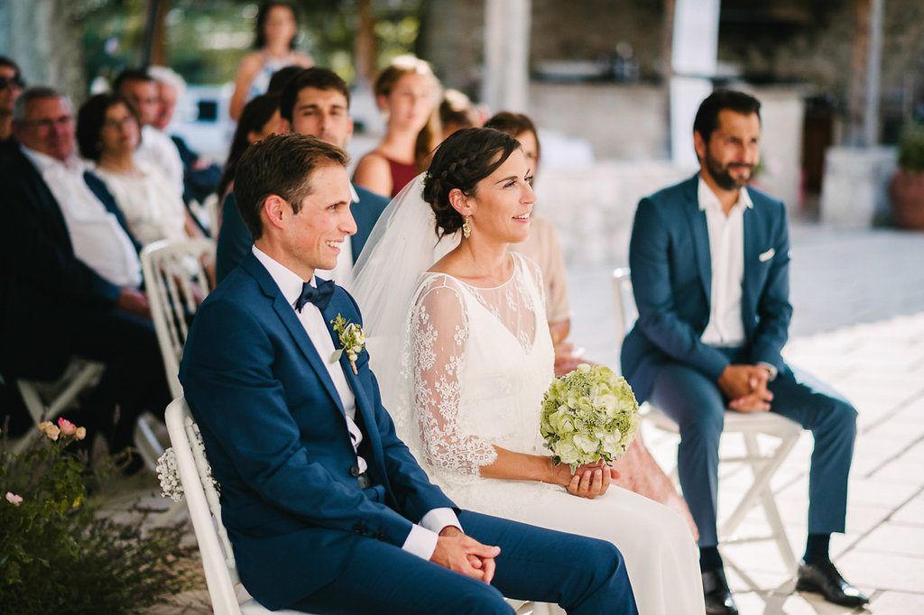 Bride and groom seated together during wedding ceremony, bride holding green and white bouquet with guests behind.