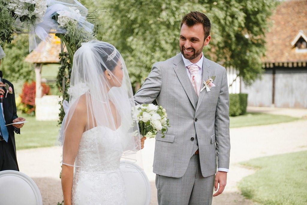 Groom in grey suit smiles at veiled bride during outdoor wedding ceremony beside rustic building.