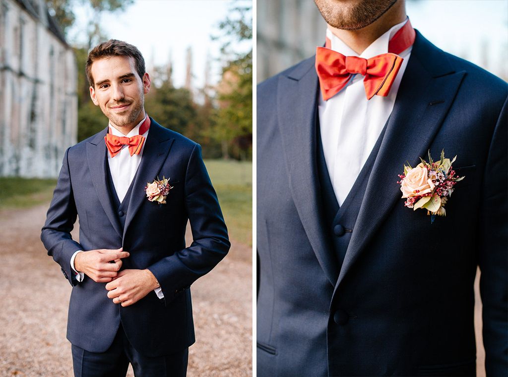 Groom in navy suit with red bow tie and floral boutonniere poses for wedding portrait.