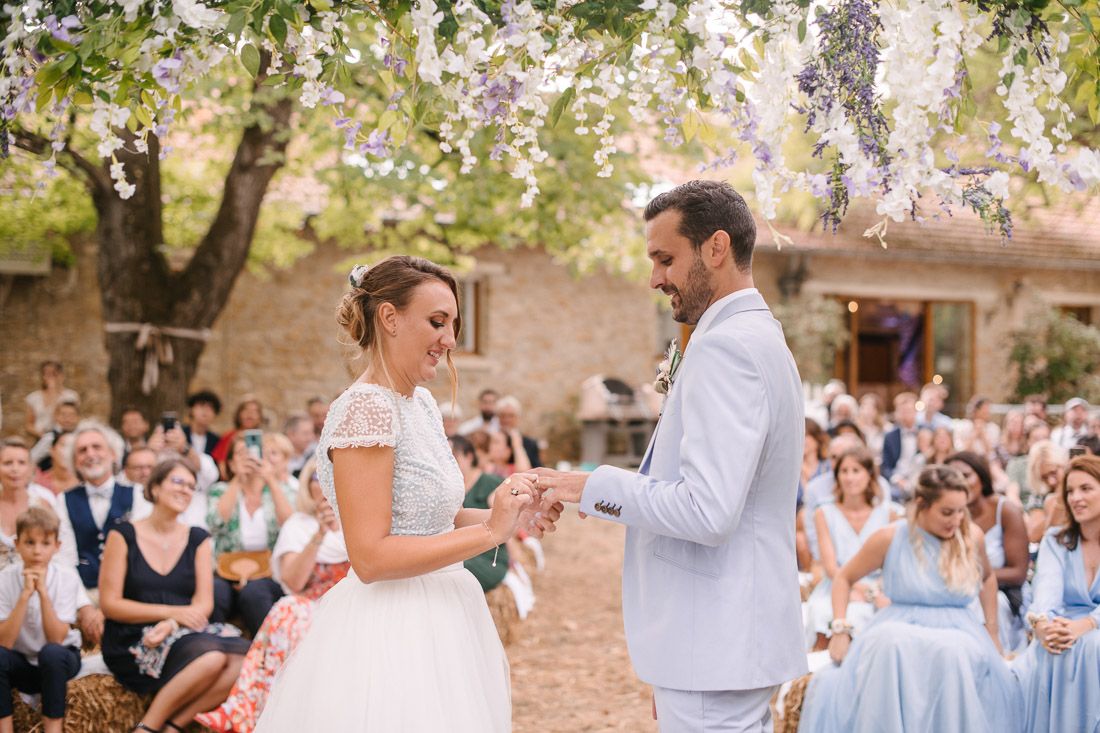 Bride and groom exchange rings during outdoor wedding ceremony under blooming wisteria tree with guests seated behind.