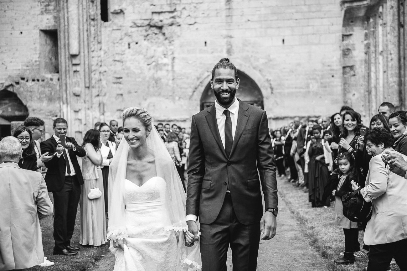 Black and white wedding photo of a tall groom and bride walking hand in hand through a cheering crowd outdoors.