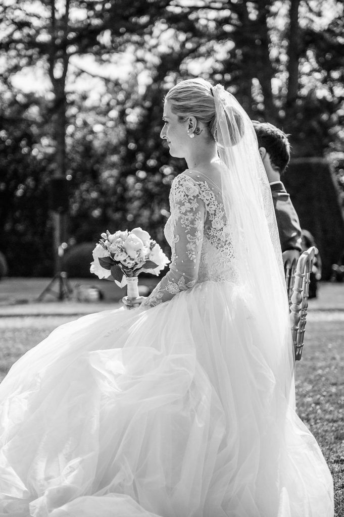 Elegant bride in lace long-sleeve gown holds floral bouquet during outdoor garden wedding ceremony in black and white.
