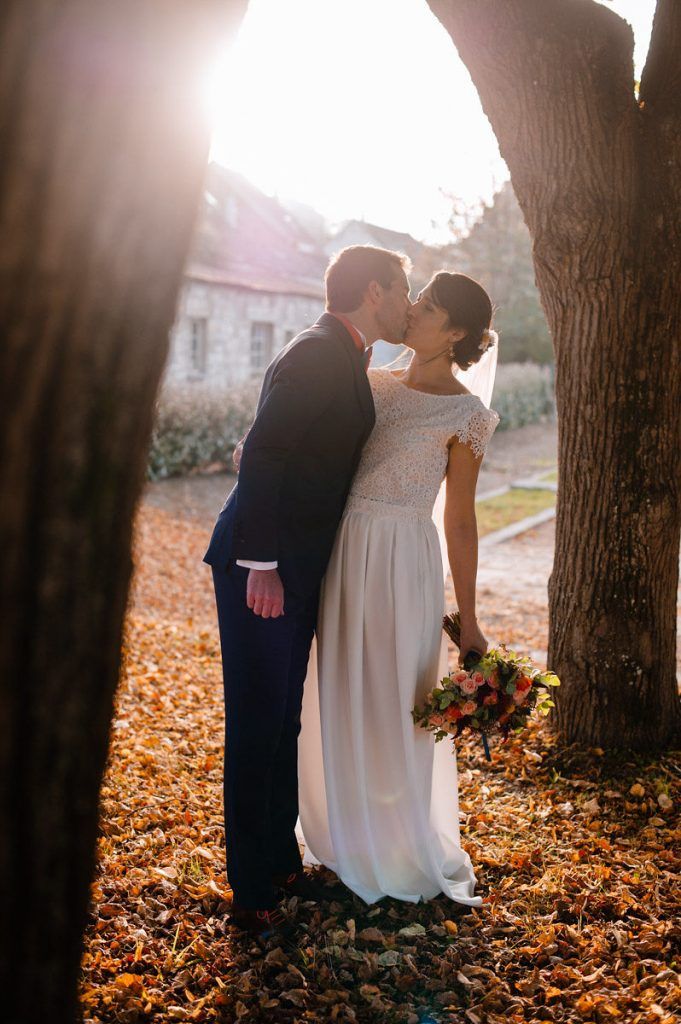 Bride and groom kiss in golden sunset light between ancient trees on autumn leaves.