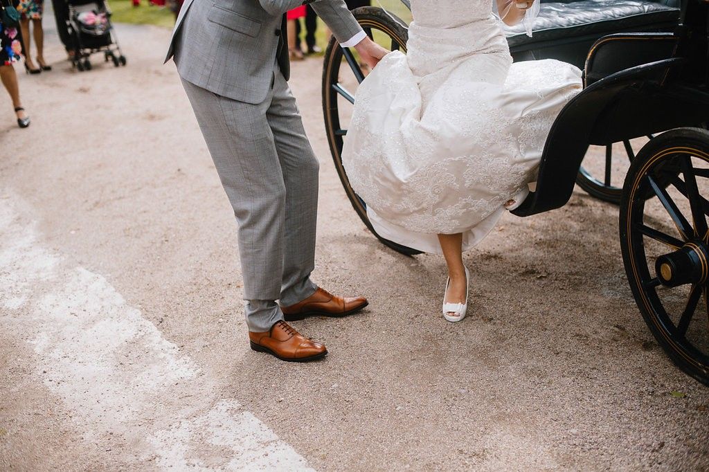 Groom helps bride in white wedding gown step down from a horse-drawn carriage, showing elegant heels.