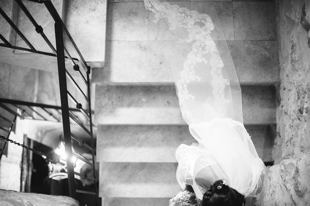 Dramatic black and white photo of bride's veil billowing on stone staircase with bouquet.