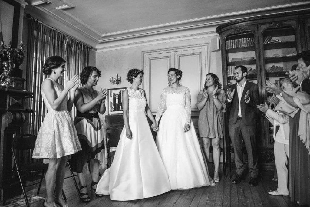 Two brides in white gowns hold hands as guests applaud during a joyful same-sex wedding celebration.
