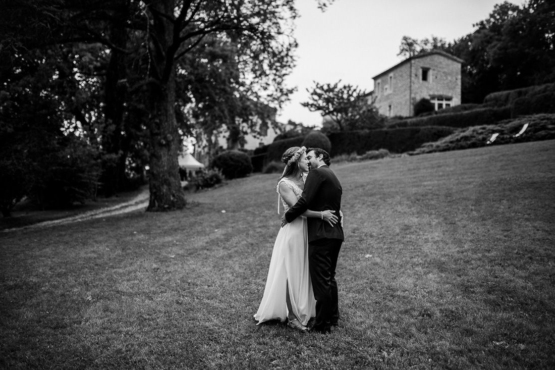 Black and white photo of bride and groom sharing a kiss on a green lawn near a stone building.