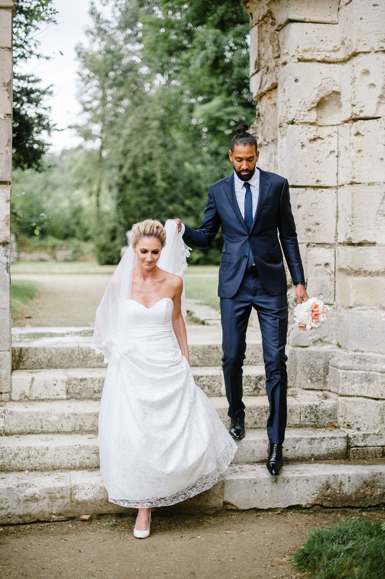 Bride in white strapless gown and veil walks down stone steps with groom in navy suit holding her veil outdoors.