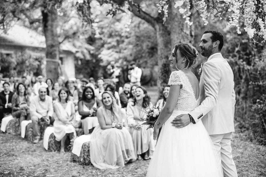 Black and white photo of bride and groom standing before smiling wedding guests outdoors.