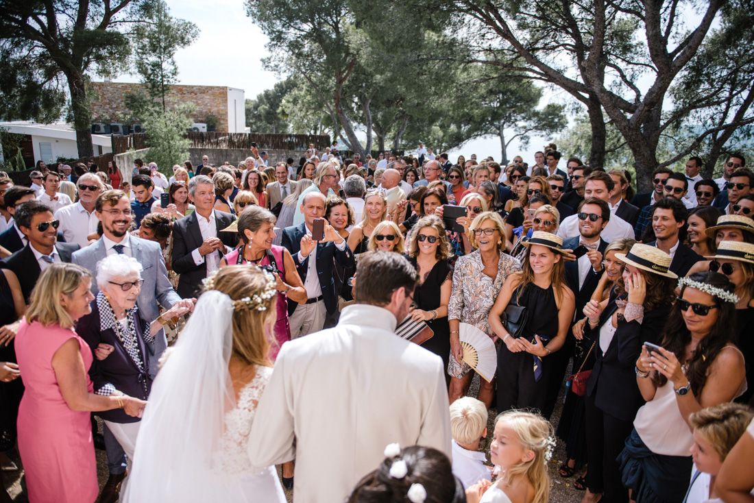 Bride and groom greeted by large crowd of wedding guests outdoors under sunny skies.