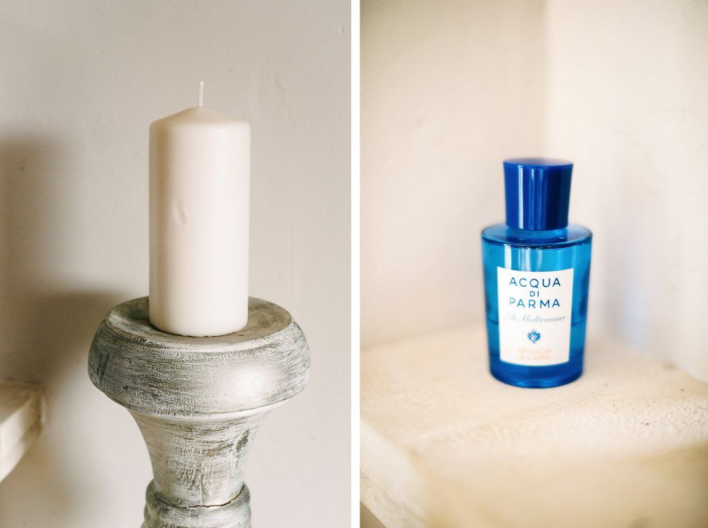 White pillar candle on rustic candleholder beside Acqua di Parma blue perfume bottle.