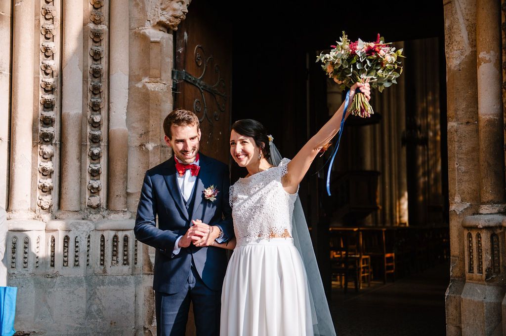 Jubilant bride raises her colorful bouquet while standing with groom at historic church entrance.