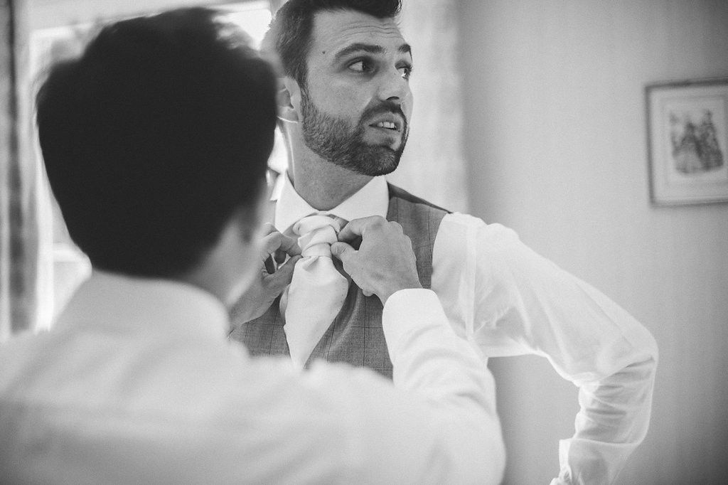 Black and white photo of groomsman adjusting groom's bow tie before wedding ceremony.