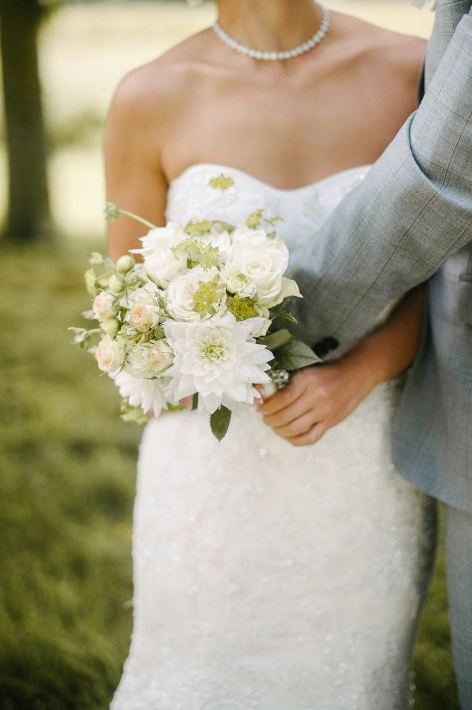 Close-up of a bride holding a white dahlia and rose bouquet while wearing a pearl necklace and lace gown.