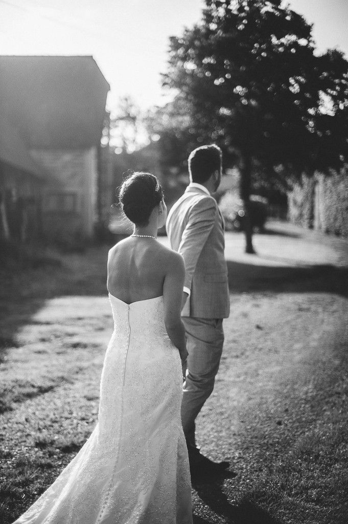 Black and white wedding photo of bride and groom walking hand in hand along a sunlit path.