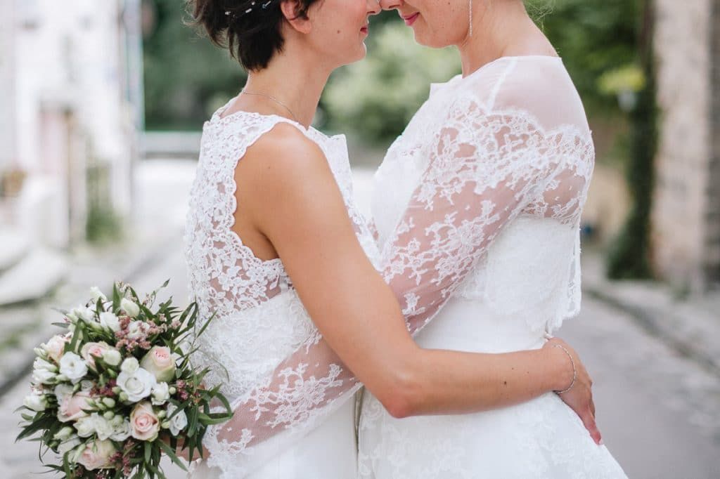 Two brides in white lace wedding gowns embrace closely while one holds a soft pink and white floral bouquet.