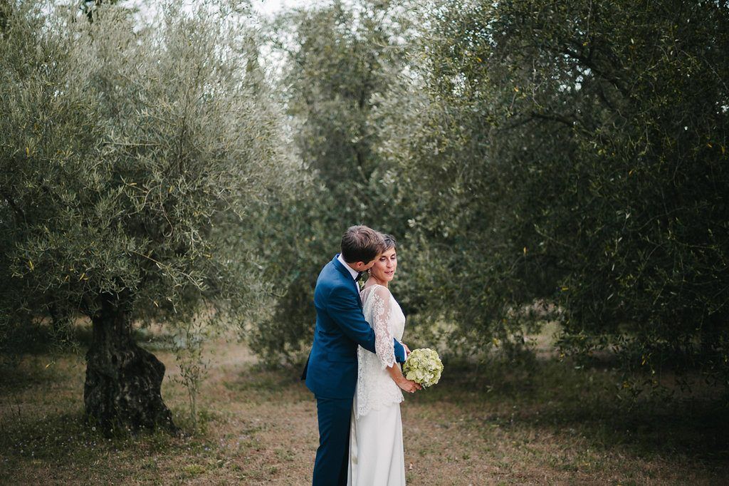Bride and groom share a romantic embrace among olive trees in a lush outdoor garden setting.