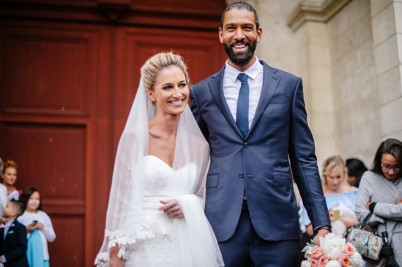 Happy newlyweds smiling outside church, bride in white gown and groom in navy suit.