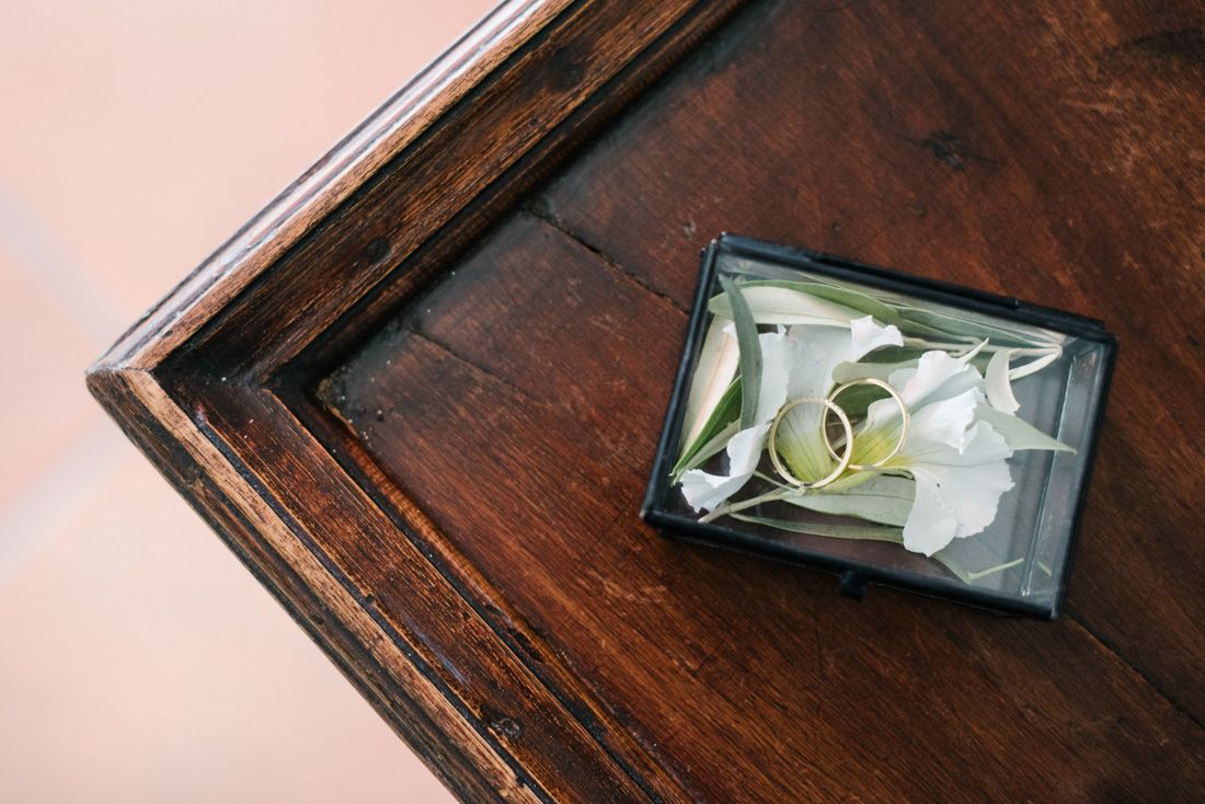 Two gold wedding rings resting on white flower petals in a small glass box on a dark wooden surface.
