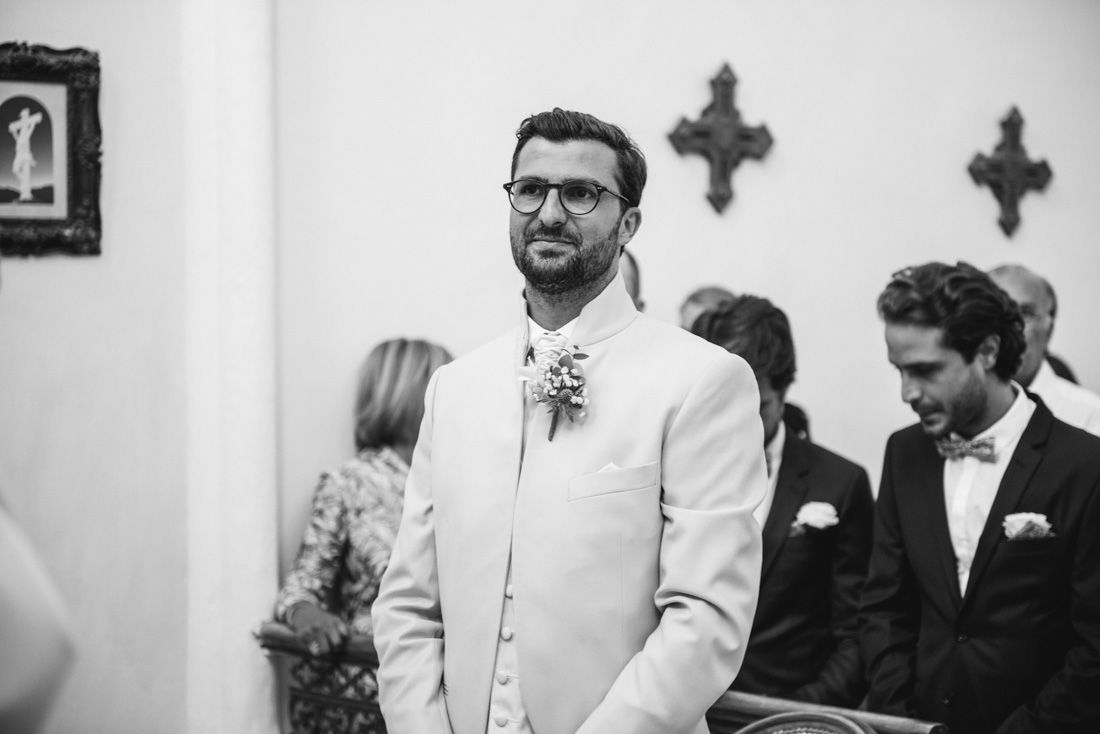 Groom in white suit smiling at altar during church wedding ceremony, black and white photo.