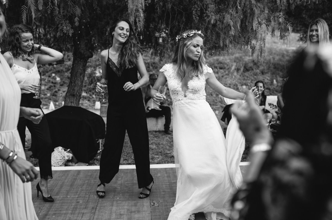 Bride in white gown dancing joyfully with a guest in black jumpsuit at outdoor wedding.