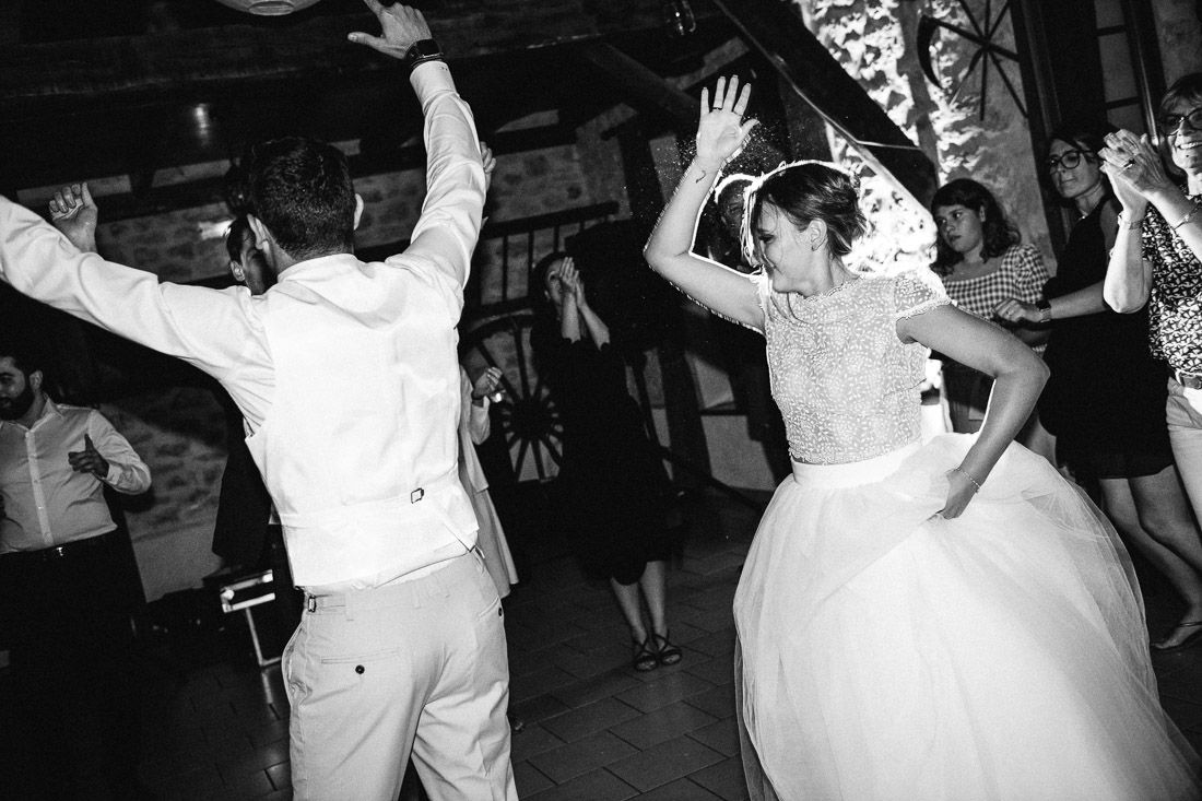 Black and white photo of bride and groom dancing energetically with guests at lively wedding reception.