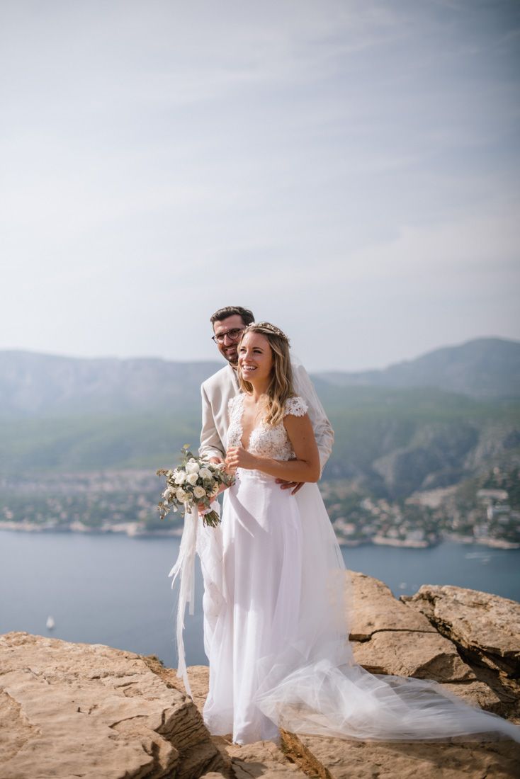 Bride and groom pose on rocky cliff with stunning mountain and bay backdrop.