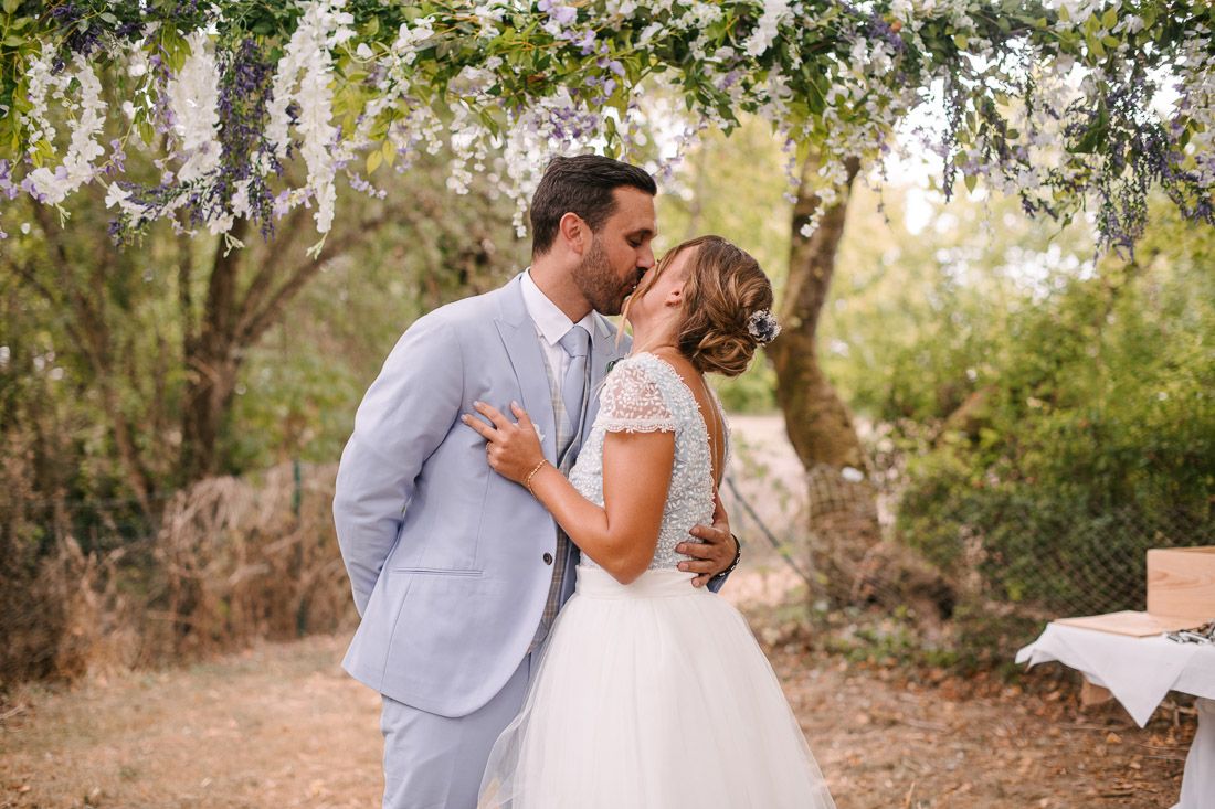 Bride and groom sharing first kiss under floral arch at romantic outdoor woodland wedding ceremony.
