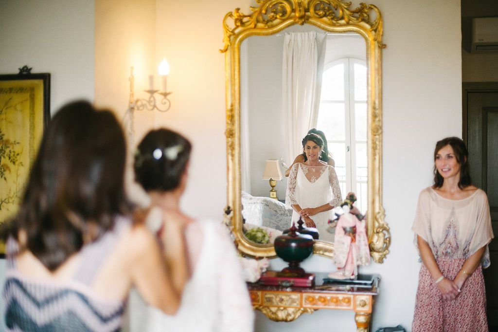 Bride in lace wedding dress viewing reflection in ornate gold mirror with smiling bridesmaid.