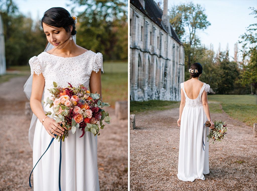 Bride in lace wedding gown holds colorful bouquet in front of historic stone building.