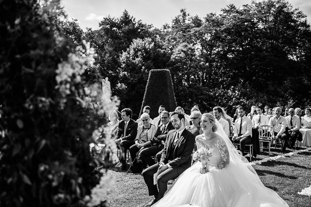 Black and white photo of bride and groom seated at outdoor garden wedding ceremony with guests and floral arch.