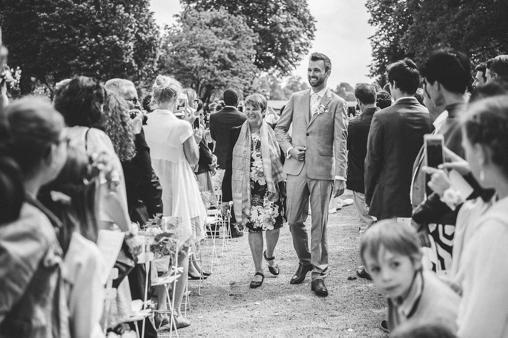 Groom escorts woman down outdoor wedding aisle lined with seated guests in black and white photo.