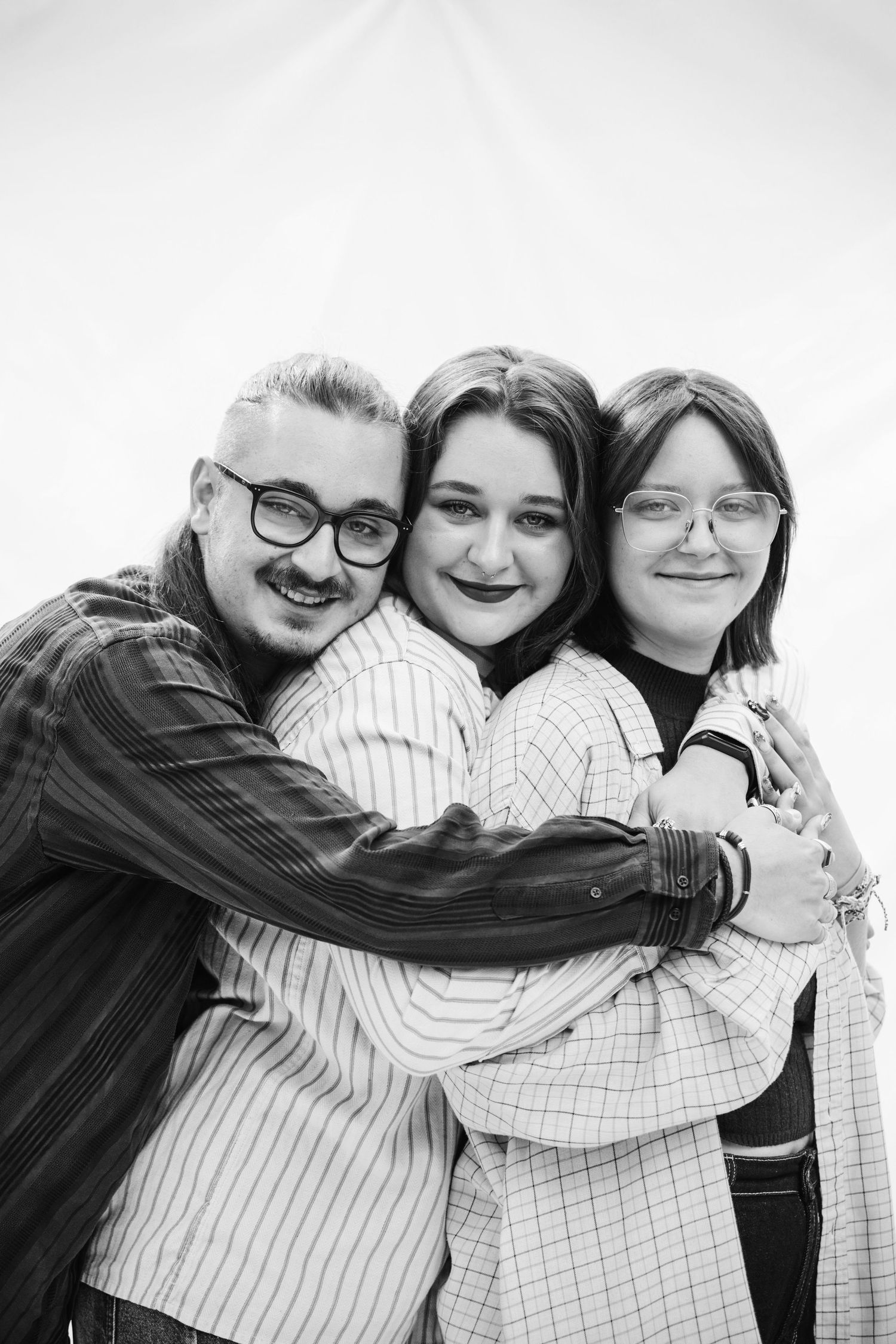 Black and white photo of three smiling young adults wearing glasses hugging each other closely.