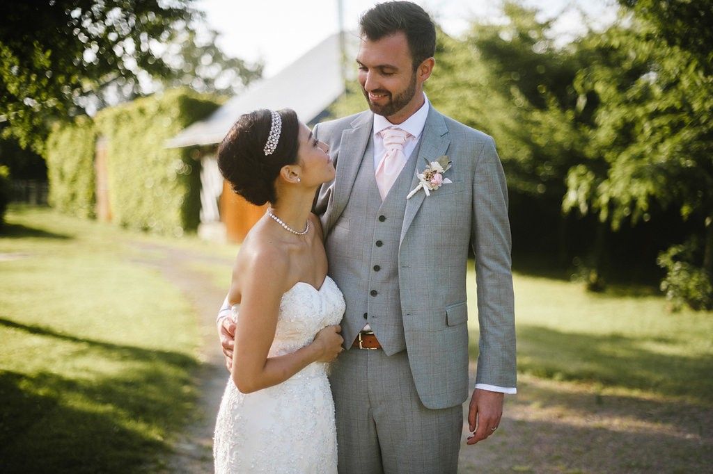 Smiling bride looks up at her groom in a grey three-piece suit with a pink boutonniere.