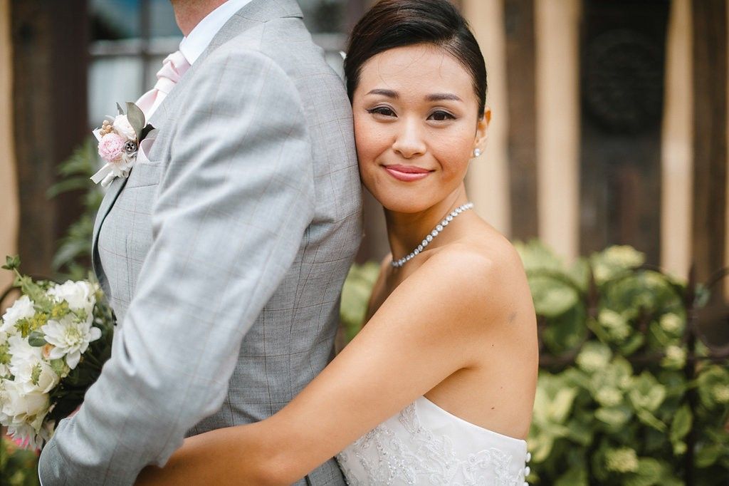 Close-up of smiling Asian bride in strapless lace gown with pearl necklace hugging groom in gray suit.