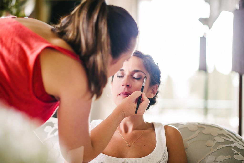 Makeup artist applying eye makeup to bride wearing white dress before wedding ceremony.