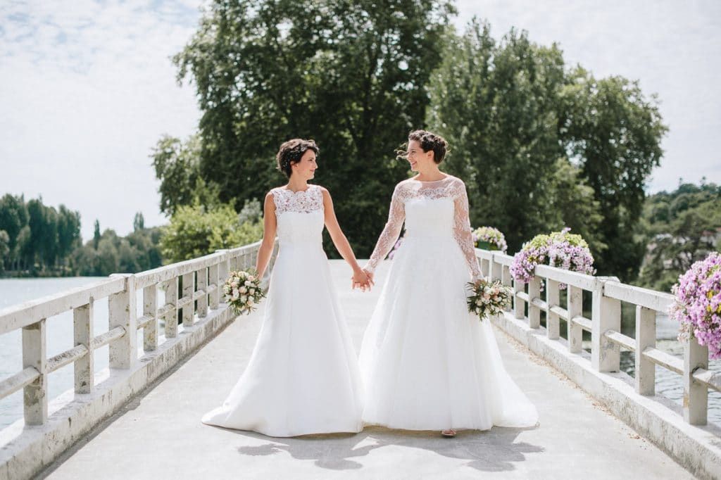 Two brides in elegant white dresses holding hands on a sunlit bridge.