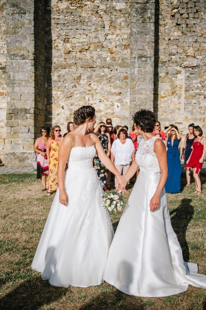 Two brides holding hands face each other before a stone wall with wedding guests behind them.