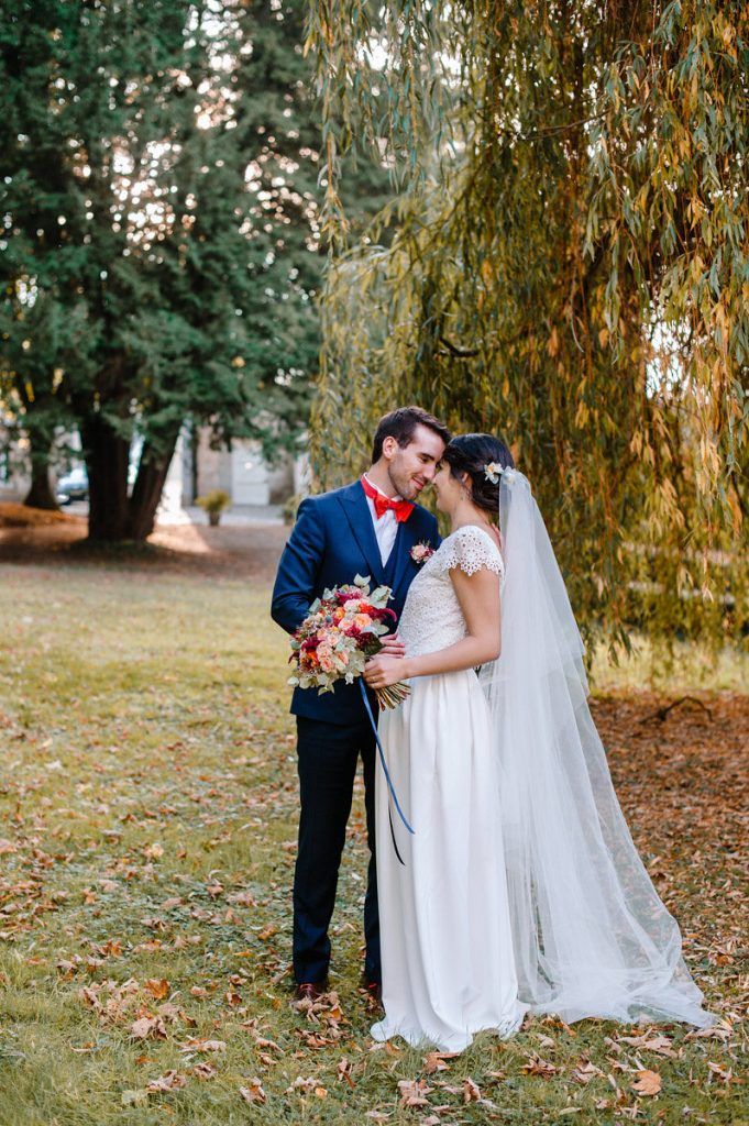 Bride and groom share tender moment under weeping willow tree in autumn park setting.