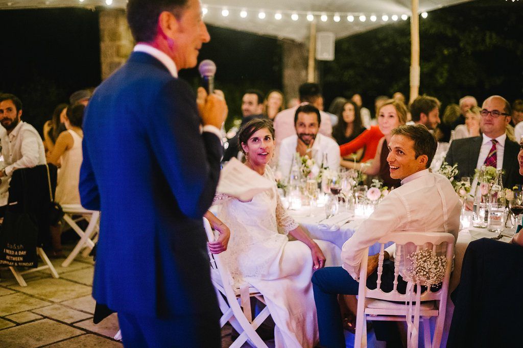 Wedding couple seated at outdoor reception table listening to guest give speech at night.
