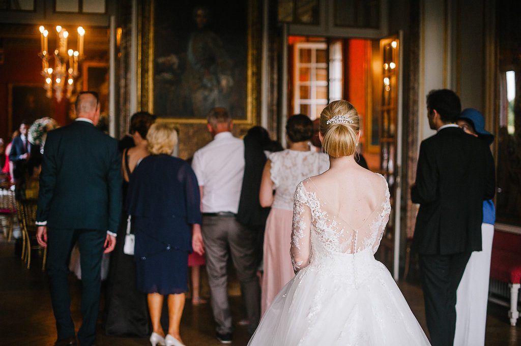 Bride in lace gown walks with wedding guests through ornate chandelier-lit chateau reception hall.