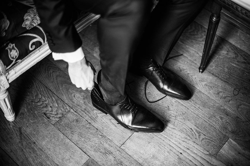 Black and white close-up of groom tying dress shoes on wooden floor before wedding ceremony.