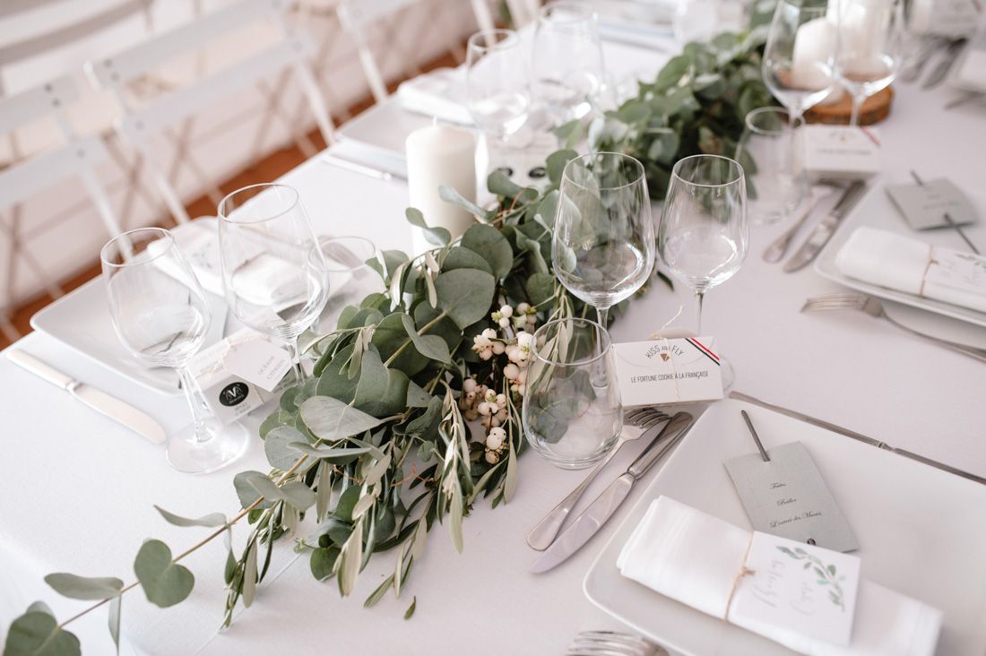 Elegant wedding table setting with eucalyptus greenery runner, white candles, and crystal glassware on white linen.