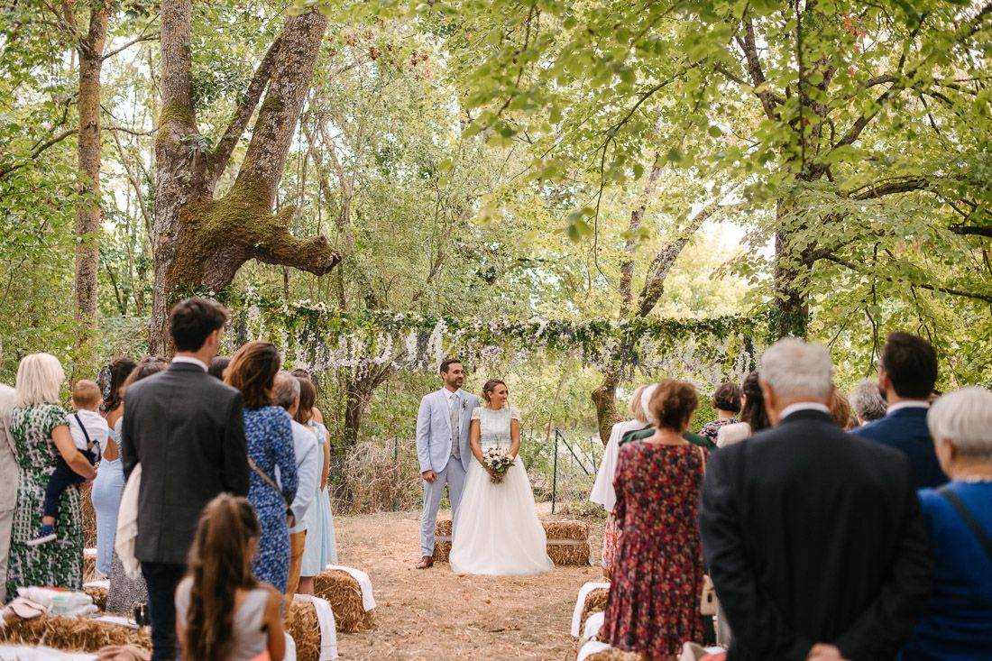 Outdoor woodland wedding ceremony with bride and groom at altar surrounded by lush trees and guests seated on hay bales.