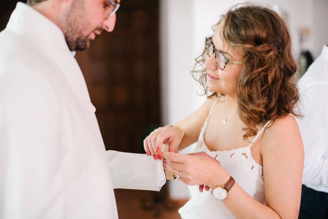 A bride in a white dress adjusts the cufflink on a groom's white suit sleeve during their wedding ceremony.