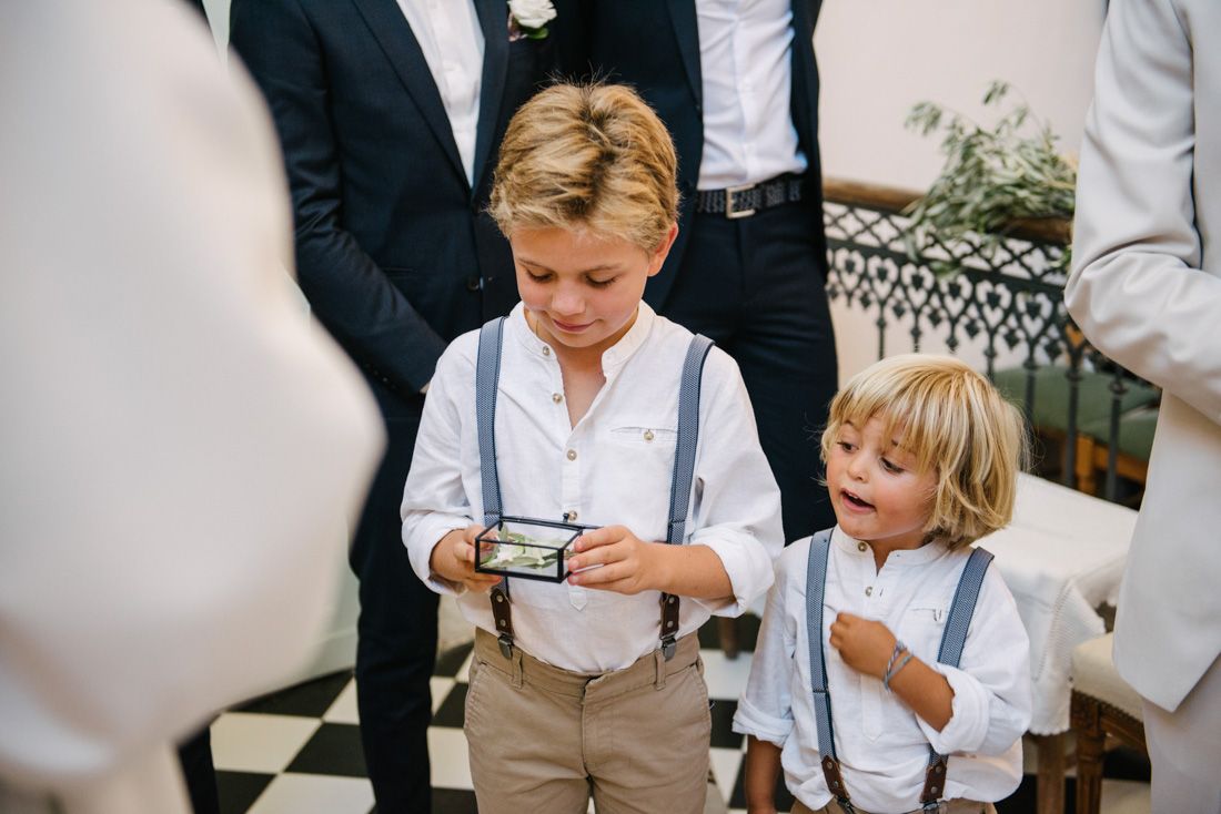 Two young ring bearers in white shirts and suspenders admire wedding rings during a ceremony.