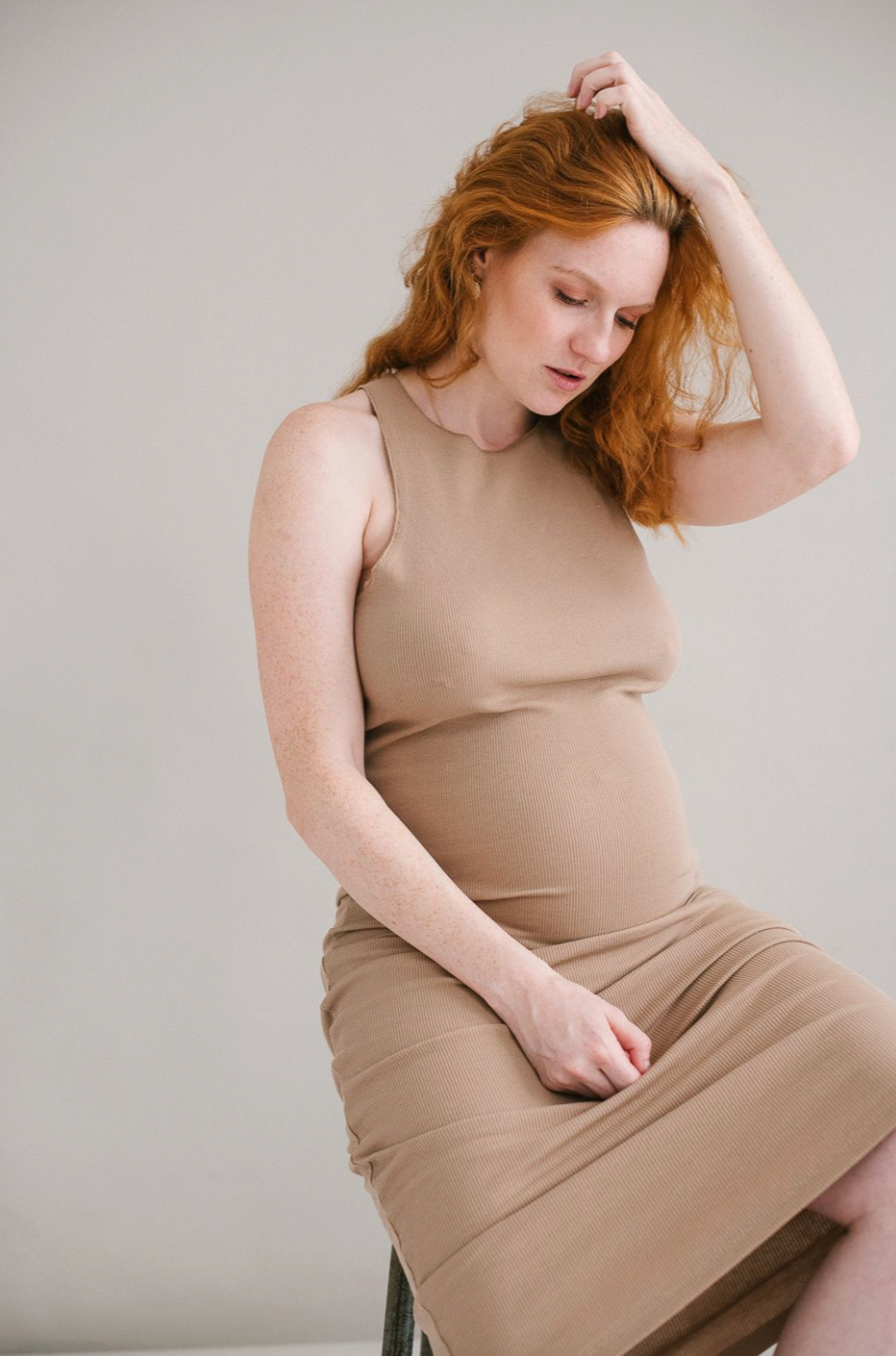 Red-haired pregnant woman in tan dress sitting gracefully with hand in hair in a studio.