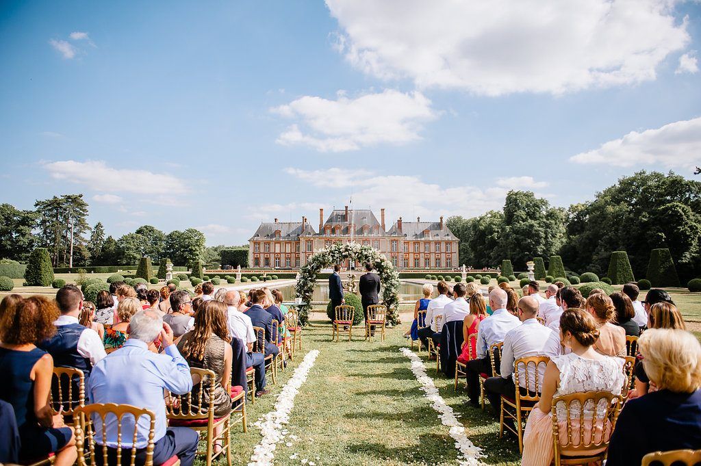 Outdoor wedding ceremony at grand French chateau with guests seated on lawn under blue sky.