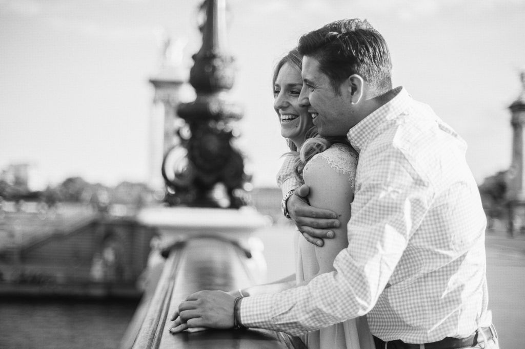 Black and white photo of a laughing couple embracing on Pont Alexandre III in Paris.